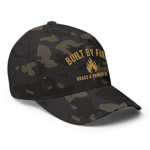 A structured baseball cap with a black and olive green camouflage pattern, featuring a gold flame logo and the text 'BUILT BY FIRE' and 'BRASS & BUNNER CO.' on the front panel.