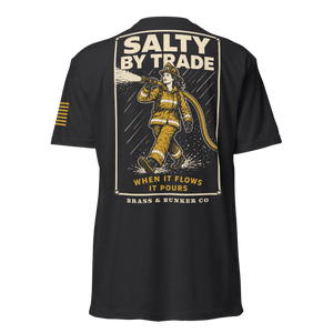 Black short-sleeve t-shirt shown from the back, featuring a large graphic print of a firefighter in yellow turnout gear spraying water, with the text 'SALTY BY TRADE', 'WHEN IT FLOWS IT POURS', and 'BRASS & BUNKER CO'. A small yellow striped graphic is visible on the right sleeve.