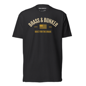 A black short-sleeve t-shirt with a gold graphic print on the back. The graphic features the text 'BRASS & BUNKER' in an arched font, a small American flag, 'ESTD 2025', and 'BUILT FOR THE BRAVE' below it.