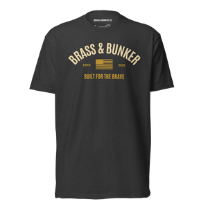 A dark charcoal gray crewneck t-shirt with a gold graphic print on the front. The graphic features the text 'BRASS & BUNKER' in an arched font, a small gold American flag, 'ESTD 2025', and 'BUILT FOR THE BRAVE' underneath.