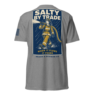 A heather gray short-sleeve t-shirt shown from the back, featuring a large graphic print of a firefighter in yellow gear spraying water under a blue background with the text 'SALTY BY TRADE' and 'BRASS & BUNKER CO'.