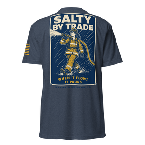 A navy blue short-sleeve t-shirt shown from the back, featuring a large graphic print of a firefighter in yellow gear spraying water, with the text 'SALTY BY TRADE' and 'WHEN IT FLOWS IT POURS'. A small yellow stripe graphic is visible on the right sleeve.