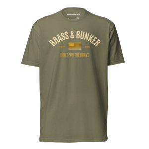 An olive green short-sleeve t-shirt with a vintage-style graphic on the chest reading 'BRASS & BUNKER' in cream and yellow, featuring a small American flag and 'BUILT FOR THE BRAVE' text below.