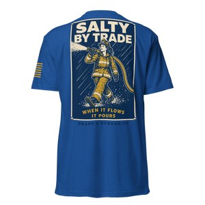 A royal blue short-sleeve t-shirt shown from the back, featuring a large graphic print of a firefighter in yellow gear spraying water under the text 'SALTY BY TRADE' and 'WHEN IT FLOWS IT POURS'. The shirt also has three gold stripes on the left sleeve.
