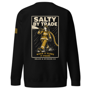 Black crewneck sweatshirt shown from the back, featuring a large graphic print of a firefighter in yellow turnout gear with the text 'SALTY BY TRADE' and 'WHEN IT FLOWS IT POURS'. A small yellow striped detail is visible on the left sleeve.