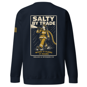 A navy blue crewneck sweatshirt shown from the back, featuring a large graphic print of a firefighter in yellow and white with the text 'SALTY BY TRADE WHEN IT FLOWS IT POURS' and 'BRASS & BUNKER CO'. A small yellow stripe detail is visible on the left sleeve.