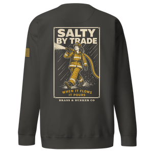 A dark charcoal gray crewneck sweatshirt shown from the back, featuring a large graphic print of a firefighter in yellow turnout gear with the text 'SALTY BY TRADE' and 'WHEN IT FLOWS IT POURS'. A small yellow striped accent is visible on the left sleeve.