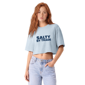 A woman with red hair wearing a light blue cropped t-shirt with 'SALTY BY TRADE' printed in navy blue on the chest, paired with light-wash blue jeans, against a plain white background.